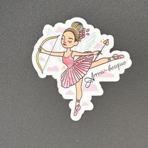May include: A sticker featuring a ballerina with a bow and arrow. The ballerina is wearing a pink tutu and pointe shoes, with a gold bow and arrow. The sticker has the text "Arrow-besque".