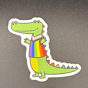 May include: A green alligator sticker with a rainbow vest and a white outline. The alligator has a smiling expression and outstretched arms. The sticker is on a dark background.