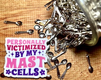 May include: A square sticker with the text "PERSONALLY VICTIMIZED BY MY MAST CELLS" in pink and purple, surrounded by small silver spoon charms spilling out of a clear container. The sticker features star and flower accents.