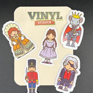 May include: A collection of vinyl stickers featuring cartoon characters from a story. The stickers include a princess in a purple dress, a soldier, a mouse king, and a man with an eyepatch. The stickers are on a beige background with the words "VINYL STICKER".