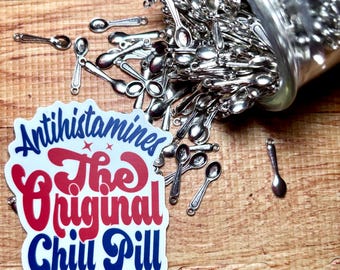 May include: A clear glass jar spilling out numerous small silver-toned metal spoon charms. Next to the jar is a white sticker with blue and red text that reads Antihistamines The Original Chill Pill in a stylized font.