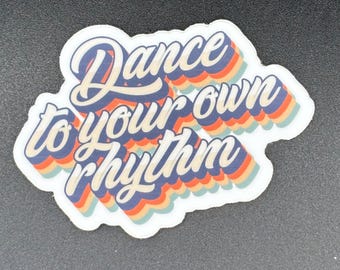 May include: A sticker with the phrase "Dance to your own rhythm" in a retro font. The text is in white with a blue outline and a layered orange, red, and teal shadow effect. The sticker has a white border.