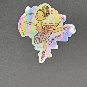 May include: Holographic sticker featuring a ballerina with a bow and arrow. The illustration shows a ballerina in a pink tutu and pointe shoes, with a bow and arrow. The text "Arrow-besque" is written in cursive. The sticker has a rainbow sheen.