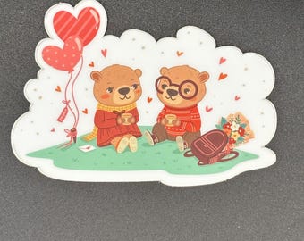 May include: A sticker featuring two cartoon bears in sweaters, sitting on grass with a picnic basket and heart-shaped balloons. The bears are holding mugs, with a bouquet of flowers and a picnic blanket nearby. The sticker has a white border.