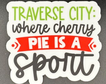 May include: A white die-cut sticker featuring the green text TRAVERSE CITY: and black cursive text where cherry. A red horizontal banner contains the white text PIE IS A, followed by the black cursive word sport at the bottom of the sticker.