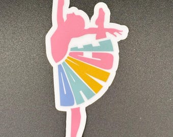 May include: A sticker featuring a pink ballerina in a graceful pose. The ballerina's tutu is a colorful design with blue, yellow, and teal stripes. The word "DANCE" is integrated into the tutu design. The sticker has a white border.