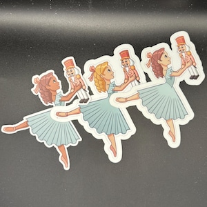 May include: Four stickers featuring a ballerina in a light blue dress holding a toy soldier nutcracker. The ballerina has brown or blonde hair and a pink bow. The nutcracker is in a red and gold uniform.