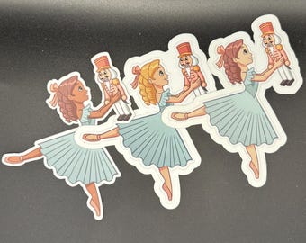 May include: Four stickers featuring a ballerina in a light blue dress holding a toy soldier nutcracker. The ballerina has brown or blonde hair and a pink bow. The nutcracker is in a red and gold uniform.