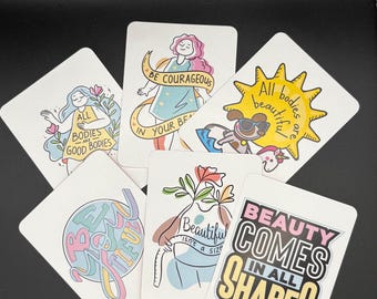 May include: A collection of white cards with colorful illustrations and text. The cards feature phrases like "All bodies are good bodies," "Be courageous," and "Beauty comes in all shapes." The illustrations include flowers, figures, and a unicorn.