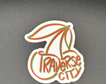 May include: A white sticker with a cherry design. The cherries are outlined in red and green, with the text "Traverse City" in green. The sticker has a glossy finish and is on a dark gray background.