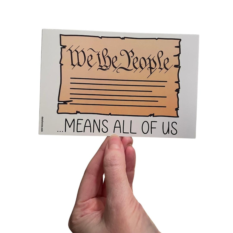 Puede incluir: Una pegatina rectangular con las palabras "We the People" en letras negras ornamentadas sobre un fondo de pergamino marr&oacute;n con l&iacute;neas horizontales. Debajo, el texto dice "...MEANS ALL OF US" en negro.