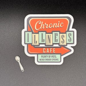 May include: A sticker with the text "Chronic Illness Cafe" in a retro diner-style design, featuring an arrow and the phrase "Plenty of Pots, Never Enough Spoons." A small silver spoon charm is also present.