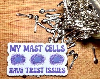 May include: A rectangular sticker with a white background and purple text reading MY MAST CELLS HAVE TRUST ISSUES, featuring three purple cell illustrations, surrounded by a pile of small silver-colored metal spoon charms.