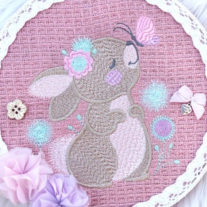 Puede incluir: Un aro de arpillera rosa con un borde de encaje blanco presenta un conejo marrón bordado con detalles rosas. El conejo lleva una corona de flores y tiene una mariposa en la cabeza. El aro está decorado con flores rosas y azules.