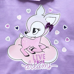 Könnte beinhalten: Lilafarbenes T-Shirt mit einem rosa Reh auf einer Wolke. Das Reh trägt eine Schleife auf dem Kopf und der Text "Live your dreams" befindet sich darunter.