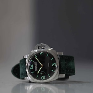 May include: A stainless steel wristwatch with a black face and green luminous numerals. The watch has a dark green fabric band. The watch face displays the brand name "Luminor Panerai" and the year "1950".