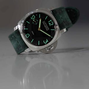 May include: A stainless steel Panerai Luminor 1950 watch with a black dial and green canvas strap. The watch features luminous hands and numerals, with the brand name and model number visible on the dial. The watch is angled slightly, showcasing its design.