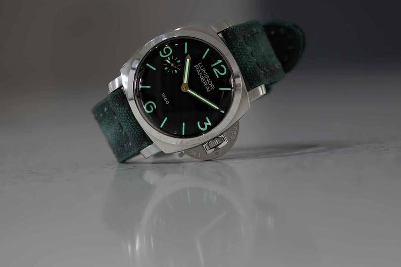 May include: A stainless steel Panerai Luminor 1950 watch with a black dial and green canvas strap. The watch features luminous hands and numerals, with the brand name and model number visible on the dial. The watch is angled slightly, showcasing its design.