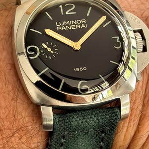May include: A close-up of a luxury wristwatch with a black face, gold hands, and a silver case. The watch has the text "LUMINOR PANERAI 1950" on the face and a dark green fabric band.