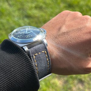 May include: A stainless steel wristwatch with a dark blue face and a grey canvas strap with yellow stitching. The watch is on a wrist, with a black long-sleeved top visible.
