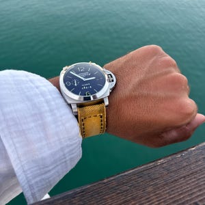 Pode incluir: Um relógio de pulso em aço inoxidável com mostrador preto, numerais brancos e uma proteção de coroa proeminente. O mostrador exibe o texto Luminor Panerai e 10 Days. Possui uma pulseira de lona texturizada em amarelo mostarda com costuras.