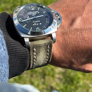 May include: A stainless steel Luminor Panerai GMT watch with a black dial and green canvas strap. The watch face displays the number 12, luminous markers, and the text "10 DAYS". The watch is secured on a wrist.