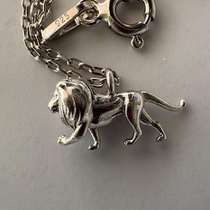 May include: A silver-coloured necklace featuring a three-dimensional lion pendant in a walking pose. The metal chain includes a circular spring ring clasp with a small rectangular tag stamped with the number 925, indicating sterling silver composition.
