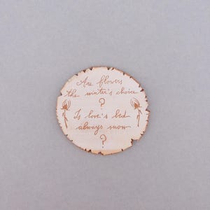 May include: A round, light brown wooden plaque with a rough edge. The plaque has handwritten text in brown ink that reads, "Are flowers the winter's choice? Is love's bed always snow?" with decorative floral elements.