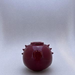 Könnte beinhalten: Eine kleine, runde, burgunderfarbene Vase mit glänzendem Finish. Die Vase hat mehrere kleine, spitze Vorsprünge um ihren Körper. Die Öffnung oben ist kreisförmig.