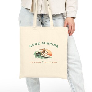 Può includere: Borsa tote color crema con la scritta "GONE SURFING" e una grafica di un surfista. La grafica mostra un surfista che cavalca un'onda con un tramonto. La borsa è realizzata in tessuto resistente e ha manici lunghi.