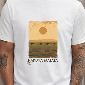 Peut inclure: T-shirt blanc avec un motif rectangulaire représentant une scène de savane avec un soleil et le texte "HAKUNA MATATA". Le motif montre un ciel jaune, de l'herbe verte et des silhouettes d'animaux.