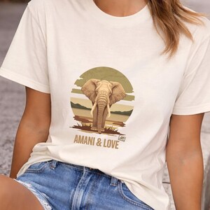 Peut inclure: T-shirt crème avec un motif d'éléphant dans un paysage de savane dans un cercle. Le texte "AMANI & LOVE" est imprimé en dessous. Le t-shirt a des manches courtes et un col rond.