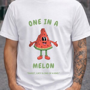 Può includere: Una t-shirt bianca a maniche corte con la grafica di una fetta di anguria in stile cartoon con braccia e gambe. Il testo sopra recita ONE IN A, e sotto recita MELON. Un testo più piccolo in basso dice "SWEET, JUICY & ONE OF A KIND."