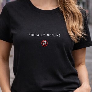 May include: A black short-sleeved t-shirt featuring the white text SOCIALLY OFFLINE centred on the chest, positioned above a small graphic of a smartphone enclosed within a red circle with a diagonal line through it, indicating a no-phone symbol.