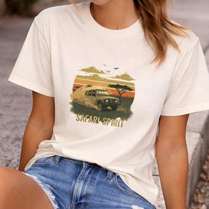 Peut inclure: T-shirt crème avec un motif de véhicule de safari dans un paysage de coucher de soleil, avec le texte "SAFARI SPIRIT" dans un style vintage. Le t-shirt est associé à un short en jean.
