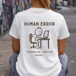 Op de afbeelding: Een wit t-shirt met een bruine afbeelding van een stokfiguur die aan een computerbureau zit, met de tekst "HUMAN ERROR" erboven en "VISIONXLAB - USER NOT FOUND." onder de illustratie, gedrukt op de achterkant van het kledingstuk.