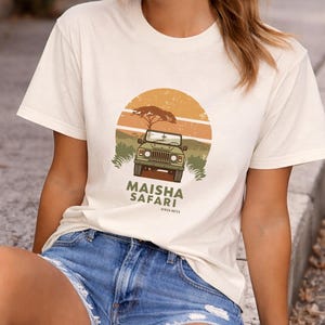 Peut inclure: T-shirt crème avec un motif d'un 4x4 de safari vert, d'un arbre et d'un coucher de soleil. Le texte "MAISHA SAFARI" est imprimé sous le véhicule. Le modèle porte un short en jean bleu.
