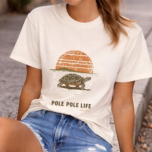 Peut inclure: T-shirt crème avec un motif de coucher de soleil, une silhouette de tortue et le texte "POLE POLE LIFE". Le t-shirt est associé à un short en jean. Le motif est imprimé dans des tons bruns et orangés.