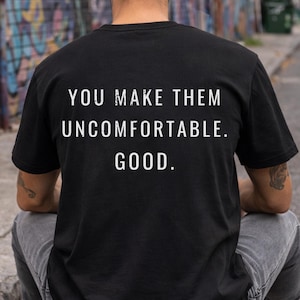 Può includere: T-shirt nero con la scritta bianca "YOU MAKE THEM UNCOMFORTABLE. GOOD.". La persona è seduta su uno skateboard. La persona indossa jeans grigi e scarpe da ginnastica bianche. Lo sfondo è un colorato muro di graffiti.