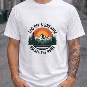 May include: White t-shirt with a circular graphic featuring a mountain scene, a setting sun, and the text "LOG OFF & BREATHE ESCAPE THE NOISE". The shirt is short-sleeved and worn by a person in dark jeans.
