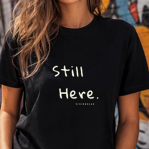 Può includere: Una t-shirt nera a maniche corte con la scritta bianca "Still Here." stampata sul petto in un carattere semplice e arrotondato, con il testo più piccolo "VISIONLAB" posizionato subito sotto in un carattere pulito e senza grazie.