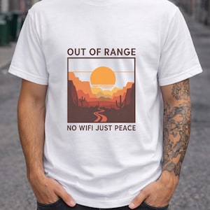 Pode incluir: Camiseta branca com um design de paisagem desértica, com um sol poente, montanhas e cactos. O texto "OUT OF RANGE" e "NO WIFI JUST PEACE" está impresso acima e abaixo do gráfico. Gola redonda e mangas curtas.