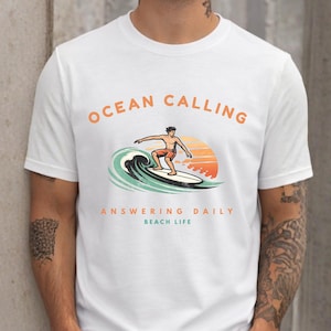 Può includere: Una t-shirt bianca a maniche corte con una grafica che raffigura una persona che fa surf su un'onda con un tramonto sullo sfondo. Il testo OCEAN CALLING, ANSWERING DAILY e BEACH LIFE è stampato in arancione e verde acqua sul petto.