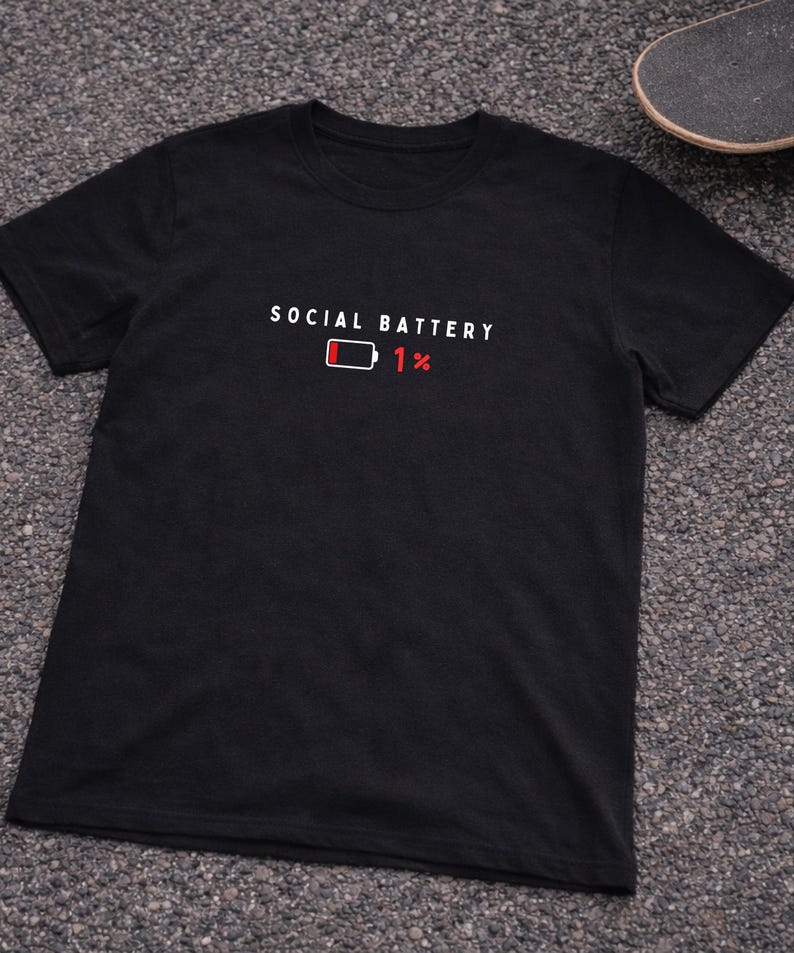 Può includere: T-shirt nera con la scritta "SOCIAL BATTERY" sopra un'icona di batteria scarica all'1%. La maglietta è realizzata in materiale morbido ed è adatta per un abbigliamento casual. Uno skateboard è sullo sfondo.