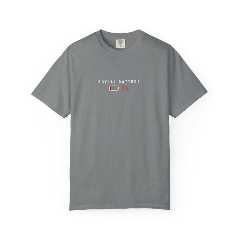 May include: A gray short-sleeved t-shirt with the words "SOCIAL BATTERY" above a graphic of a low battery indicator showing 1%. The shirt has a crew neck and a tag at the back of the neck.