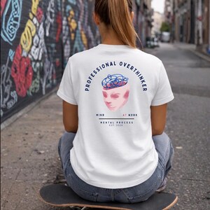 Può includere: T-shirt bianca con la scritta "PROFESSIONAL OVERTHINKER" e una grafica di una testa con un cervello aggrovigliato. La maglietta include anche le parole "MIND AT WORK" e "MENTAL PROCESS EST. 2020". La persona è seduta su uno skateboard.
