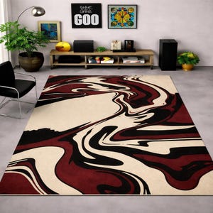 May include: A large rug featuring a cream backdrop with a swirling design in burgundy and black. The pattern resembles flowing paint, creating a modern, artistic look. The rug is placed on a grey concrete floor, suitable for a living room or similar space.