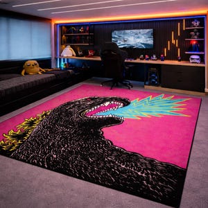May include: A pink rectangular area rug featuring a black Godzilla illustration breathing blue and yellow fire. The rug is in a bedroom with a black bed, desk, and shelves with figurines and a computer monitor.