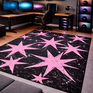 May include: A rectangular area rug with a black background and a pattern of large, pink starbursts. The stars have a hand-drawn, textured appearance. The rug is placed on a hardwood floor in a room with a desk and computer monitors.