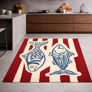Könnte beinhalten: Ein rechteckiger Teppich mit rot-weißem Streifenmuster. Zwei blaue Fischmotive sind auf dem Teppich zentriert. Der Teppich liegt auf einem grauen Fliesenboden in einer Küche.